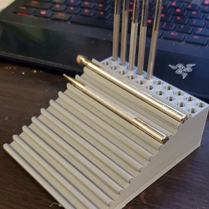 Puede incluir: Un organizador de herramientas de plástico gris con múltiples ranuras para guardar herramientas pequeñas. El organizador tiene un diseño inclinado y está lleno de varias herramientas de metal.