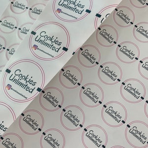 May include: A sheet of white stickers with the text "Cookies Unlimited" and the social media handle "@cookiesunlimited" printed in black and pink. The stickers are arranged in rows and columns.