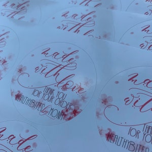 May include: Round sticker with pink watercolor floral design. The sticker says "made with love" in cursive script. Below the text is "Thank you for your order" and "HEALTHYBITES by MONA".