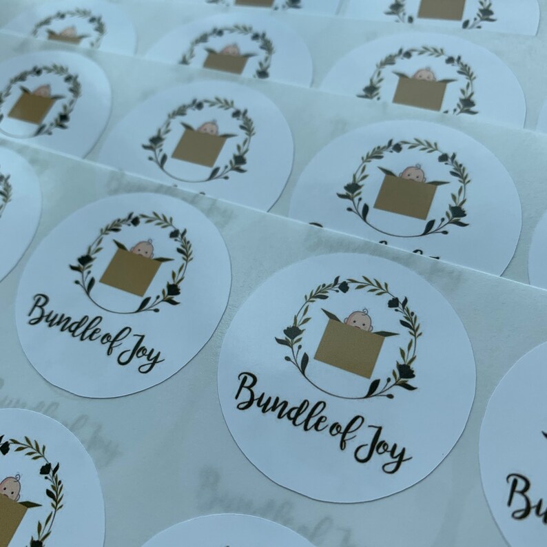 May include: A sheet of white round stickers with a black and gold design. The design features a baby inside a box with a floral wreath around it. The text "Bundle of Joy" is written in black cursive lettering.