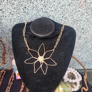 May include: A gold wire necklace with a flower pendant featuring a white stone in the center. The necklace is displayed on a black velvet bust form.