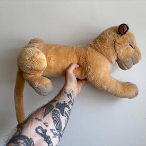 May include: A plush lioness toy in shades of tan and brown, with a long tail and detailed facial features. The stuffed animal is held up against a plain white background.