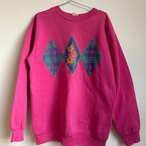 Puede incluir: Una sudadera rosa con un diseño floral en el centro. El diseño presenta tres formas de diamante con un patrón de cuadros y un motivo floral en el centro.