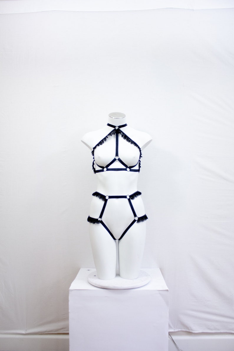 May include: A black lingerie set on a white mannequin. The set features a halter-neck bra, high-waisted briefs, and a choker, all with lace trim and ring details. The lingerie is made of a dark material, possibly satin.