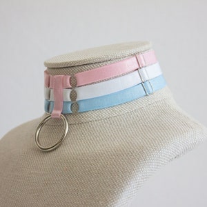 May include: A choker necklace with three bands of pink, white, and light blue fabric. The choker has a silver ring attached to it.