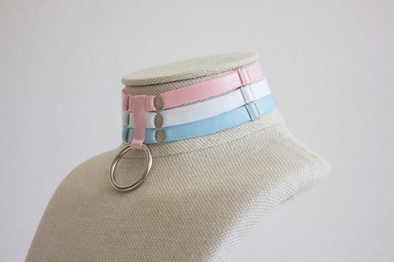 May include: A choker necklace with three bands of pink, white, and light blue fabric. The choker has a silver ring attached to it.