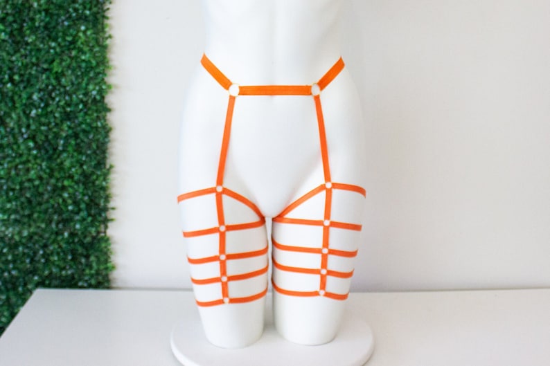 Op de afbeelding: Oranje lingerie harnas met banden en ringen. Het harnas is op een wit mannequins.