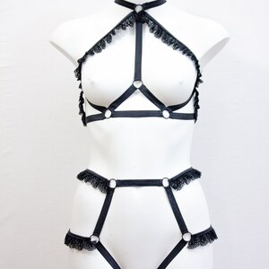 May include: Black lingerie set on a mannequin. The set comprises a halter-neck top with lace edging and a high-waisted bottom. The design incorporates straps and metal rings for a distinctive appearance.
