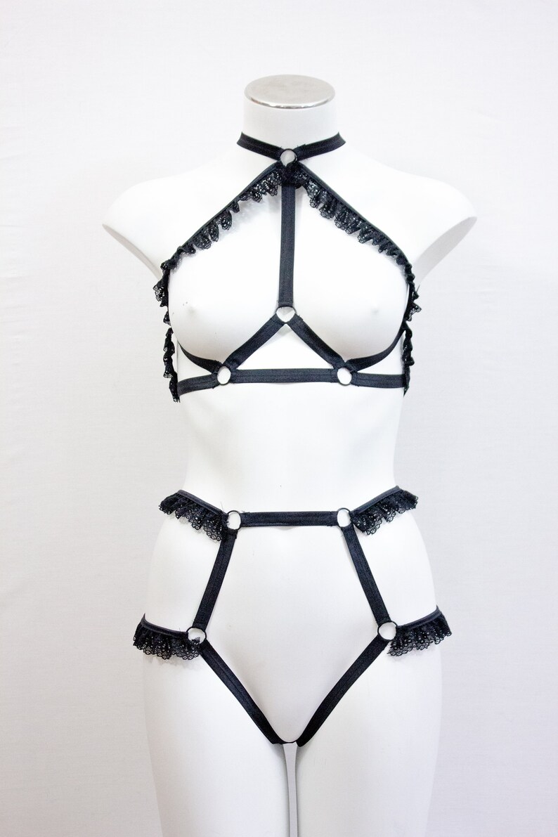 May include: Black lingerie set on a mannequin. The set comprises a halter-neck top with lace edging and a high-waisted bottom. The design incorporates straps and metal rings for a distinctive appearance.