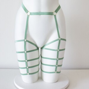 Op de afbeelding: Saliegroene kousenband met gouden ringen en banden. De kousenband is ontworpen om op de dijen te worden gedragen en heeft een brede band die om de taille gaat.