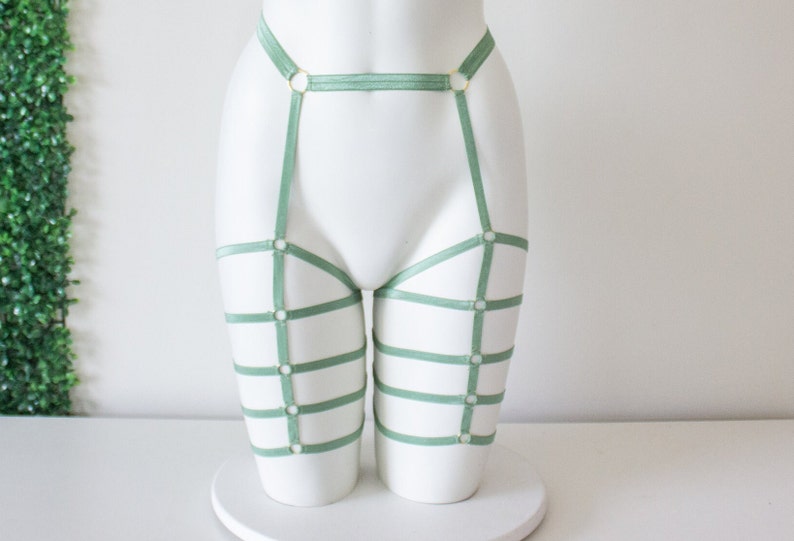 Op de afbeelding: Saliegroene kousenband met gouden ringen en banden. De kousenband is ontworpen om op de dijen te worden gedragen en heeft een brede band die om de taille gaat.