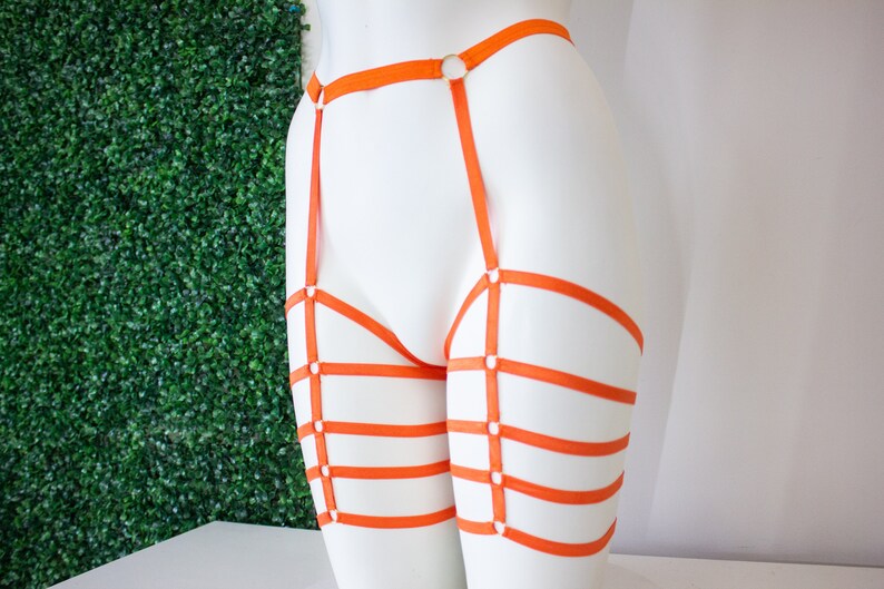 Op de afbeelding: Oranje lingerie harnas met meerdere banden en ringen. Het harnas is op een wit mannequins.