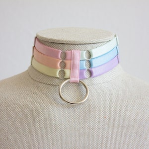 May include: A pastel-colored choker with a silver ring. The choker is made of four layers of ribbon in shades of light blue, pink, peach, and yellow.