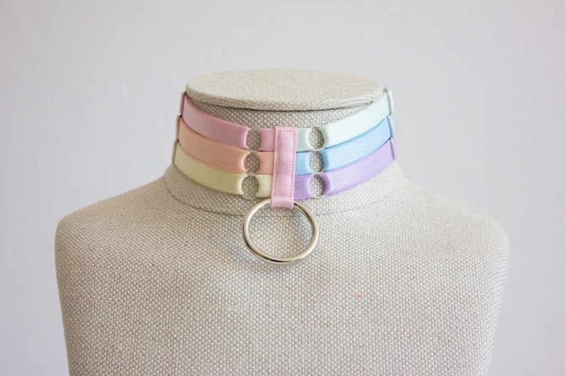 May include: A pastel-colored choker with a silver ring. The choker is made of four layers of ribbon in shades of light blue, pink, peach, and yellow.
