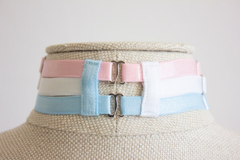 May include: A set of three adjustable bra straps in pink, white, and light blue. The straps have silver metal clasps.