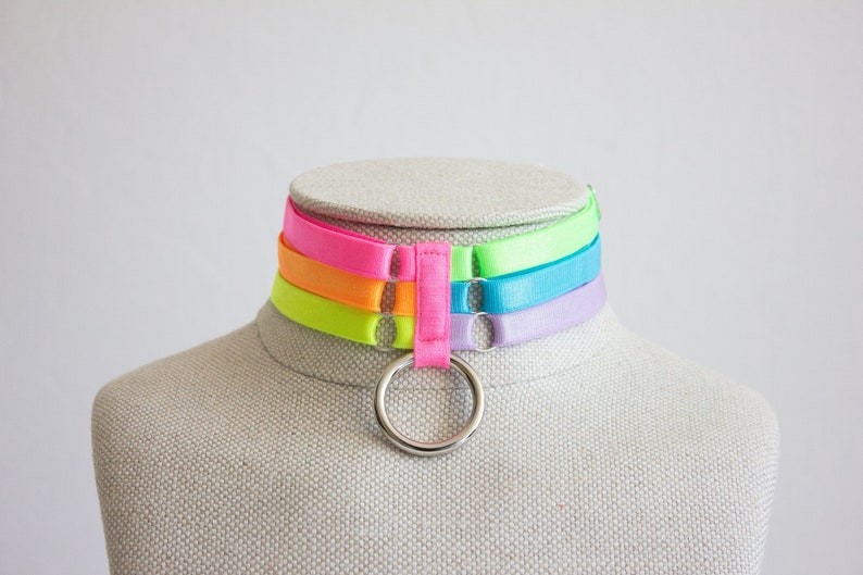May include: A multi-layered choker necklace with a silver ring. The choker is made of four layers of neon colored ribbon: green, orange, pink, and purple.