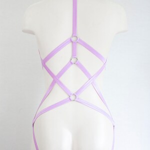May include: A lavender-colored lingerie harness with multiple straps and silver rings. The harness is designed to be worn over the shoulders and around the waist.