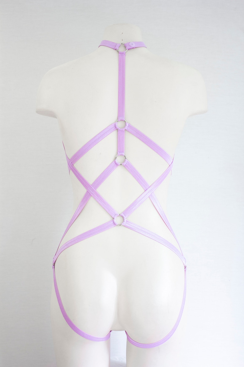 May include: A lavender-colored lingerie harness with multiple straps and silver rings. The harness is designed to be worn over the shoulders and around the waist.