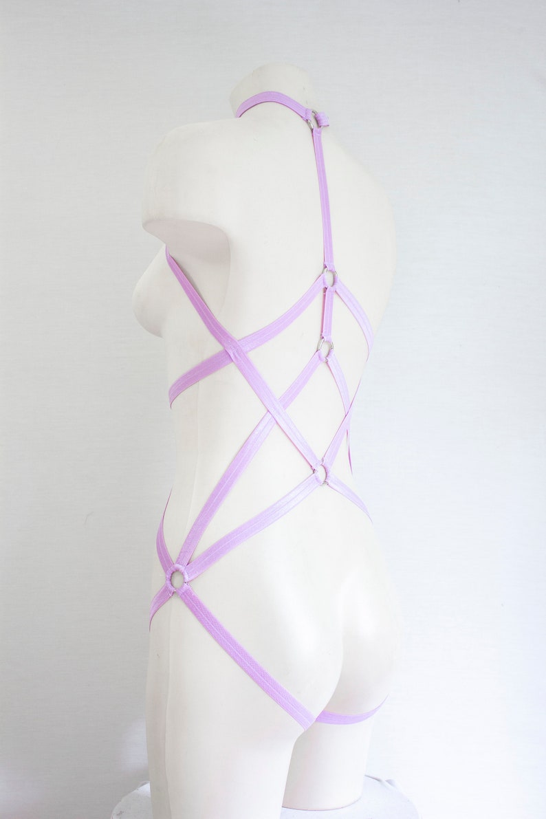 May include: A lavender colored body harness with multiple straps and silver metal rings. The harness is on a white mannequin.