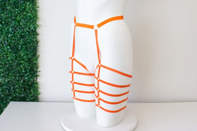 Op de afbeelding: Oranje lingerie harnas met meerdere banden en gespen. Het harnas is gemaakt van een zacht, rekbaar materiaal en is ontworpen om over de heupen en dijen te worden gedragen.