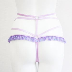 May include: A light purple lingerie set featuring a strappy design with a ruffled trim. The set includes a high-waisted harness and a thong. The ruffled trim has a glittery, iridescent effect.