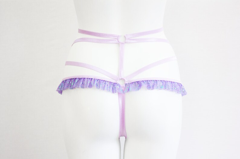 May include: A light purple lingerie set featuring a strappy design with a ruffled trim. The set includes a high-waisted harness and a thong. The ruffled trim has a glittery, iridescent effect.