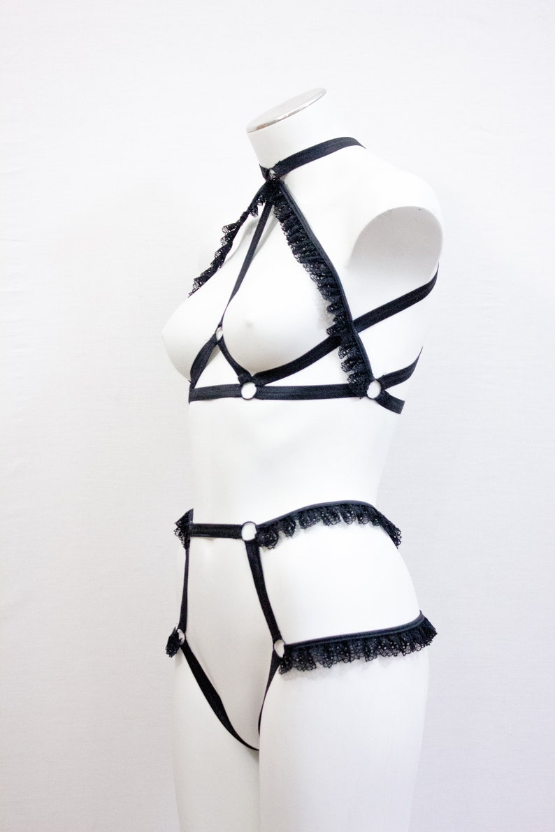 May include: Black lingerie set on a white mannequin. The set includes a halter top with lace trim and a matching high-waisted bottom. The lingerie features a strappy design with metal ring accents.