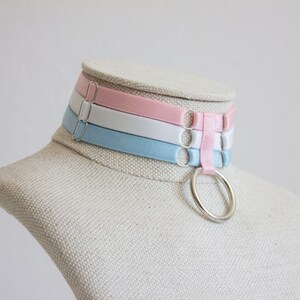 May include: A layered choker necklace with three bands of light blue, white, and pink fabric. The choker has a silver ring attached to the center band.