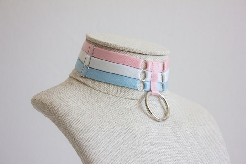 May include: A layered choker necklace with three bands of light blue, white, and pink fabric. The choker has a silver ring attached to the center band.