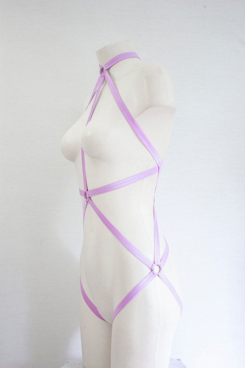 May include: A lilac-colored body harness with multiple straps and rings. The harness is made of a soft, stretchy material and is designed to be worn over the body.