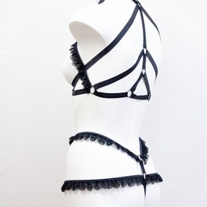 May include: Black lingerie set displayed on a white mannequin. The set features a bra with lace detailing and a harness design, a matching thong, and a garter belt. The lingerie is crafted from a soft, black material.