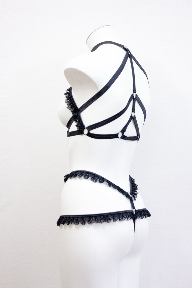 May include: Black lingerie set displayed on a white mannequin. The set features a bra with lace detailing and a harness design, a matching thong, and a garter belt. The lingerie is crafted from a soft, black material.