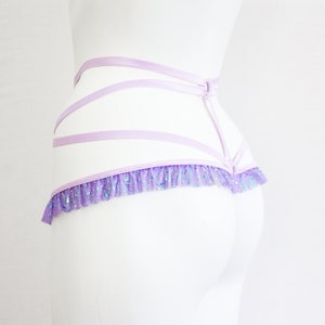 May include: A light purple lingerie set with a layered design. The set features multiple horizontal straps and a ruffled trim with iridescent glitter. The lingerie is displayed on a white mannequin against a white background.