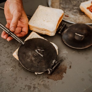 May include: A person using a cast iron sandwich press to make a grilled cheese sandwich. The press has a long handle and is being used to press down on the sandwich. The sandwich is on a grey surface.