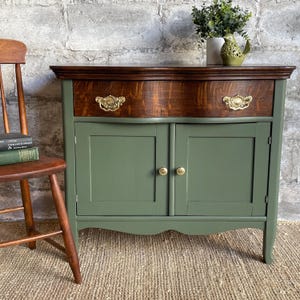 May include: A vintage wooden cabinet with a single drawer and two doors. The cabinet is painted a deep green color with brass hardware. The top of the cabinet is a rich brown wood.