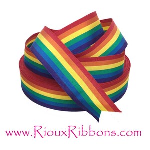 May include: A roll of rainbow-colored grosgrain ribbon. The ribbon features a vibrant spectrum of colors, including red, orange, yellow, green, blue, and purple. The ribbon is tightly wound, showcasing its texture and color variations. The website address www.RiouRibbons.com is visible.