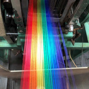 May include: A close-up of a textile machine weaving colorful threads. The threads are arranged in a rainbow spectrum, transitioning from red to violet. The machine's metal components are visible, with a focus on the weaving process.