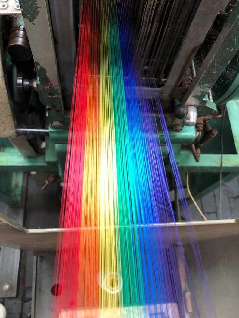 May include: A close-up of a textile machine weaving colorful threads. The threads are arranged in a rainbow spectrum, transitioning from red to violet. The machine's metal components are visible, with a focus on the weaving process.