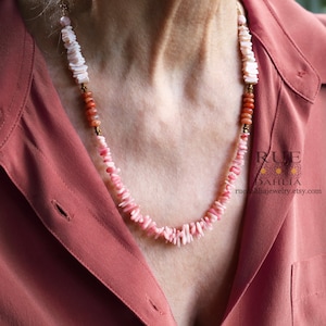 May include: A pink and white shell necklace with gold accents. The necklace is layered with different sizes of shells and is worn by a person with a red shirt.