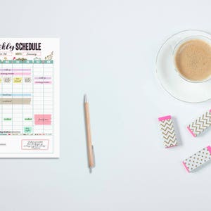 May include: A weekly schedule planner with a cup of coffee, a pen, and three erasers. The planner has a colorful layout with the words "Weekly Schedule" at the top. The erasers have a white base with gold chevron and pink accents.