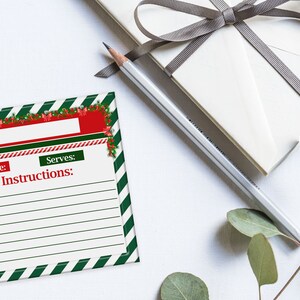 以下が含まれることがあります： クリスマスをテーマにしたレシピカード。赤と緑のボーダーがあり、材料と手順を記入するスペースがあります。鉛筆、灰色のリボンで結ばれた白い紙に包まれたギフト、ユーカリの葉も画像に写っています。