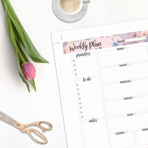 May include: A weekly planner with floral accents, a pink tulip, and gold scissors on a white surface. The planner includes sections for priorities, to-do lists, and notes, with days of the week listed.