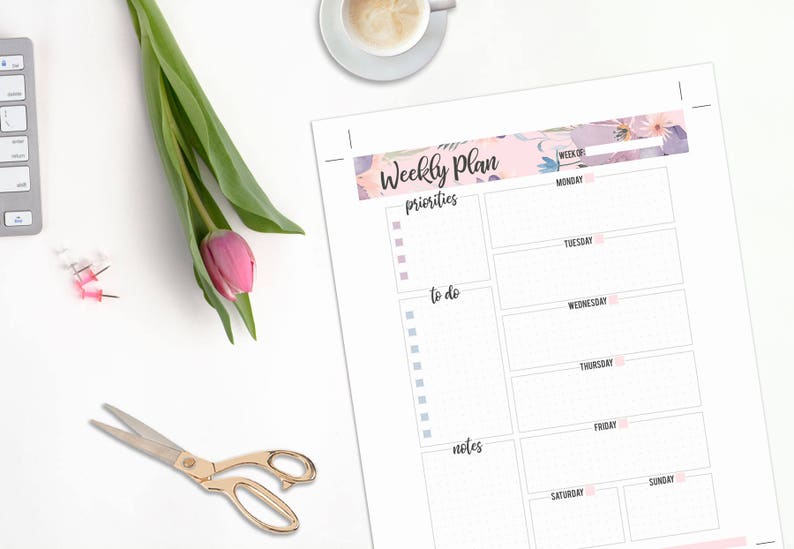 May include: A weekly planner with floral accents, a pink tulip, and gold scissors on a white surface. The planner includes sections for priorities, to-do lists, and notes, with days of the week listed.