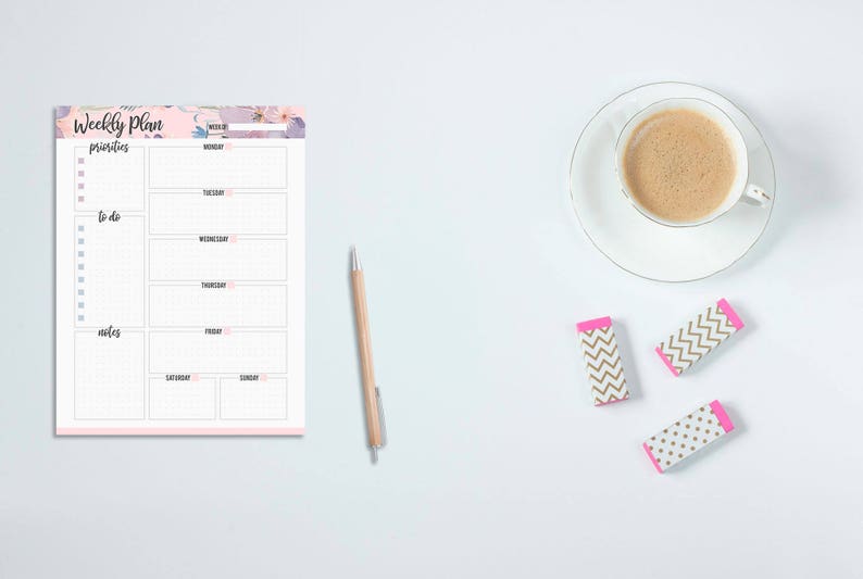 May include: A weekly planner notepad with floral accents, a pen, three erasers, and a cup of coffee on a white surface. The planner has sections for each day of the week, priorities, to-do lists, and notes.