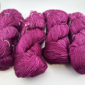 May include: Four skeins of vibrant magenta yarn arranged side by side. Each skein features a twisted, multi-ply construction with a subtle sheen and deep purple undertones visible throughout the fibre texture. A small white paper tag is attached.