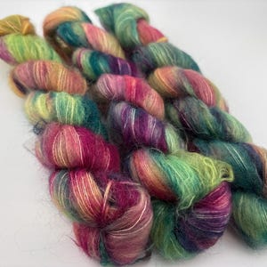 May include: Three skeins of hand-dyed yarn featuring a variegated color palette of vibrant magenta, lime green, teal, and golden yellow. The yarn has a fuzzy, mohair-like texture with a soft halo effect, twisted into loose, elongated bundles.