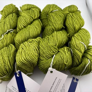 May include: Four skeins of lime green yarn arranged in a square formation. Each skein is secured with a white string and features a white paper tag with blue accents. The yarn has a twisted, multi-ply texture and a vibrant, solid colour.