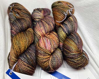 May include: Close-up of several skeins of hand-dyed yarn in shades of brown, gold, purple, and green. The yarn is tightly wound and has a textured appearance. The skeins are arranged on a white surface. A blue tag is visible.