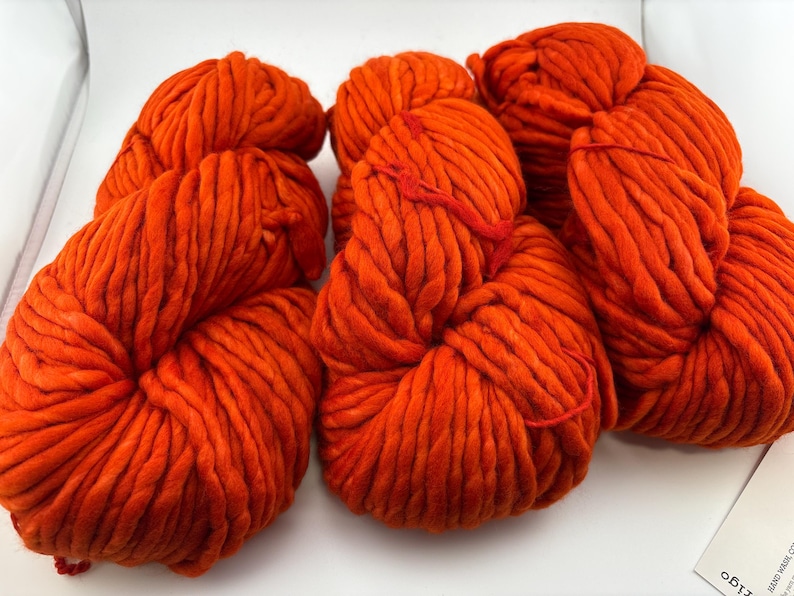 K&ouml;nnte beinhalten: Drei Str&auml;nge aus dickem, leuchtend orangefarbenem Garn, die nebeneinander angeordnet sind. Jeder Strang weist eine grobe, gedrehte Textur mit weichem Aussehen auf. Das Garn hat durchgehend eine einheitliche, helle orange Farbe.
