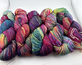 May include: Five skeins of hand-dyed yarn arranged in a row. The yarn features a variegated color palette of vibrant pink, deep purple, bright yellow, and teal blue, with each skein displaying a unique blend of these saturated hues.